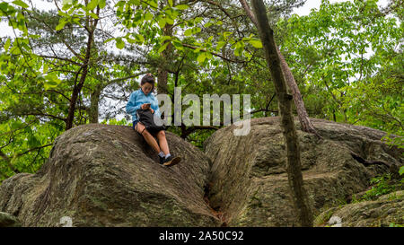 Teen girl in blue shirt and shorts sitting on large boulder looking at mobile phone Stock Photo