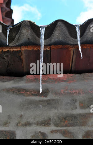 Dark roof with icicles, view from ground on top, gray brick wall, winter cloudy blue sky Stock Photo