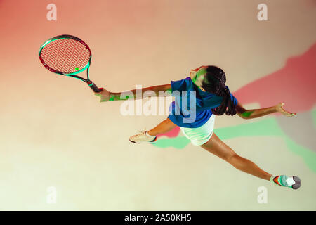 Young woman in blue shirt playing tennis. She hits the ball with a racket. Indoor studio shot with mixed light. Youth, flexibility, power and energy. Top view. Stock Photo
