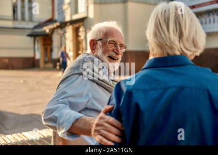 That's so funny! Happy senior bearded man in glasses looking at his wife and smiling while sitting on the bench together Stock Photo