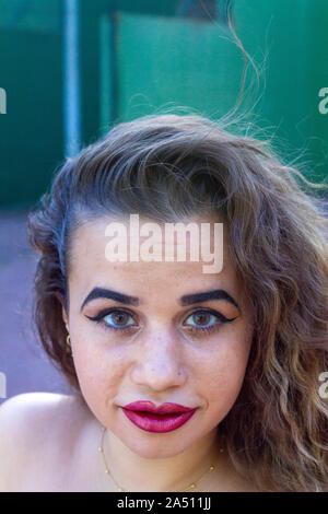 Portrait of young woman with urban clothes, with curly hair. On a green wall in the background, made up to party. Stock Photo