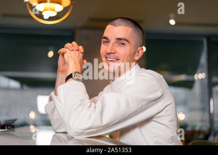 Young smiling handsome cook chief in white uniform posing in modern luxury restaurant interior. Photo with endarkened background Stock Photo