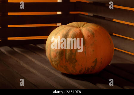 A bright orange pumpkin lies on an old, dark wooden surface against the background of the walls of wooden slats through which bright fiery light break Stock Photo