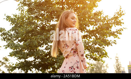nice young woman with loose fair hair in pink dress smiles and poses for camera against bright sun close view Stock Photo