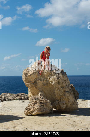 One blonde caucasian woman with red shirt sitting on a big stone in Bahrija area in Malta with blue sea in the background. Woman hiking and relaxing n Stock Photo