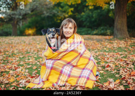Woman wrapped in woollen blanket with dog in autumn fall park. Beautiful happy young Caucasian woman sitting on ground hugging domestic canine animal Stock Photo