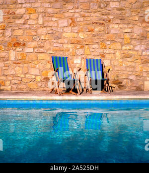 Two wooden deckchairs at the edge of a swimming pool Stock Photo