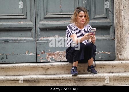 A young woman with her phone sits on the steps of a disused church in Rome, Italy Stock Photo