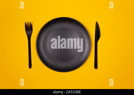 Black plastic plate, fork, knife on yellow background. Creative look at table setting. Fast food and street food concept. Stock Photo