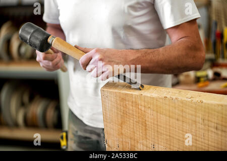 Carpenter or woodworker working with a chisel cutting or carving a block of wood with the beveled blade in a woodworking workshop in a close up on his Stock Photo