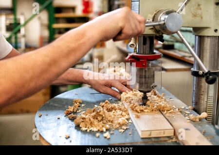 Woodworker or carpenter using a vertical drill to drill a hole in a wooden block with wood shavings in a woodworking workshop in a close up on his han Stock Photo