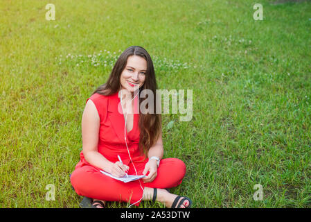 Portrait of young smiling business woman in red suit in earphones listening to music, doing online learning cources and writing in notebook while sitt Stock Photo