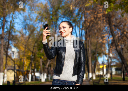 Calling by phone happy woman. Portrait of a young beautiful brunette girl in a black leather jacket on a background of autumn street Stock Photo