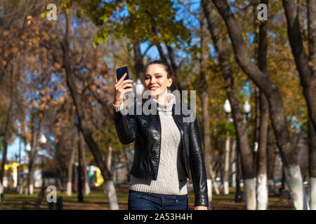 Calling by phone happy woman. Portrait of a young beautiful brunette girl in a black leather jacket on a background of autumn street Stock Photo
