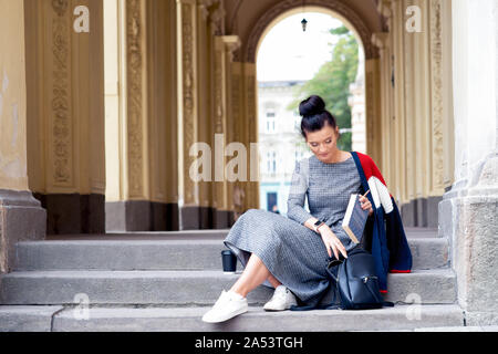 Student girl is putting the books into black backpack on stairs of university. Woman is putting a book into backpack on stairs. Education concept. Stock Photo