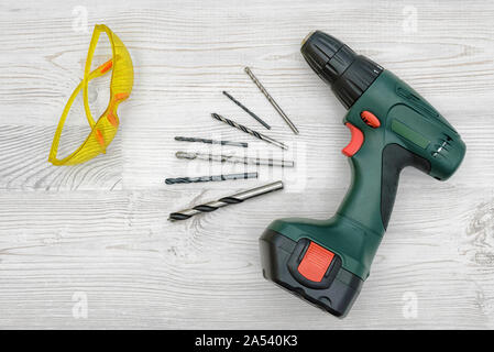 A cordless drill set on a wooden table background with a set of bits in the box and yellow protective glasses around. Stock Photo