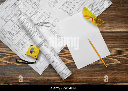 Blueprints on wooden background with piece of paper, compass, pencil, measuring tape and protective glasses. Stock Photo
