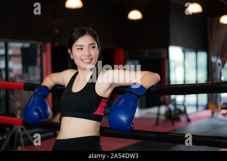 Tired Asian boxer sports girl leaned on black red ropes on boxing ring, and have a rest after hard training in black loft gym. Sporty fit for healthy Stock Photo
