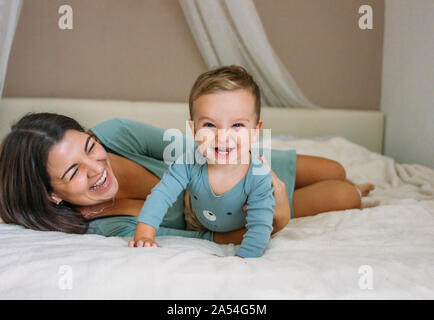 Charming happy little baby boy having fun with mom brunette woman on bed in the bright bedroom