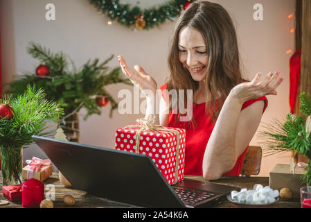 A beautiful woman in a red dress is holding a gift box and ordering online purchases on her laptop. Online shopping for Christmas holidays. Stock Photo