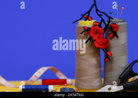 Two large spools of thread stand on a blue background. In the photo there is an application in the form of red flowers, scissors for sewing, buttons a Stock Photo