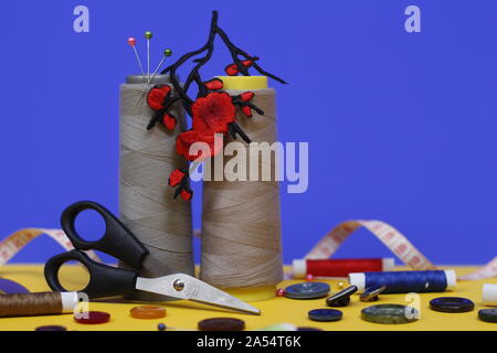 Two large spools of thread stand on a blue background. In the photo there is an application in the form of red flowers, scissors for sewing, buttons a Stock Photo
