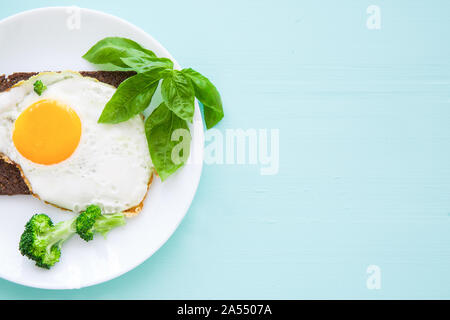 Fried eggs with fresh basil leaves, broccoli and rye bread on bright colorful turquoise backgroud. Stock Photo