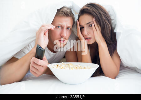 young couple handsome man and beautiful woman watching horror movie in bed eating pop corn looking frightened Stock Photo