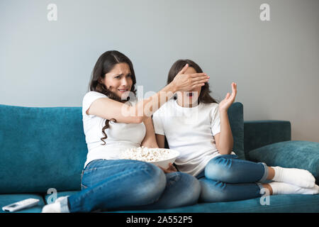 beautiful young mother with her cute teenager daughter watching scary movie on the sofa eating popcorn looking frightened Stock Photo