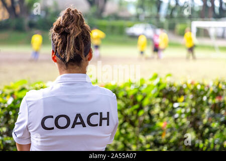 Back view of a female soccer, football, coach in white coach shirt watching her team play at an outdoor football field Stock Photo