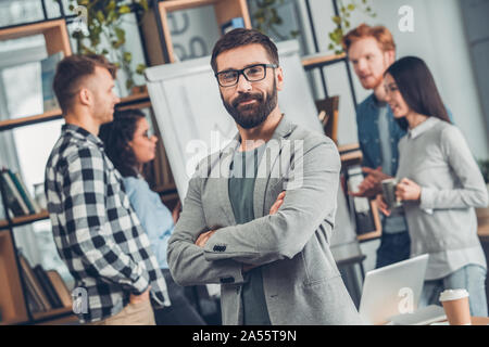 Startupers working together at office standing bearded man in glasses close-up looking camera smiling successful while colleagues discussing project Stock Photo