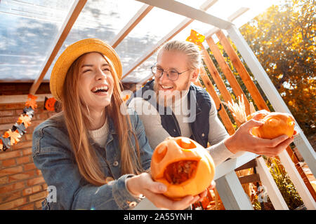 Halloween Preparaton Concept. Young couple leaning on fence decorating house with jack-o'-lantern with carved pumpkins laughing cheerful side view Stock Photo