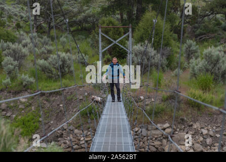 Man In Blue Shirt Stands On Suspension Bridge across river Stock Photo