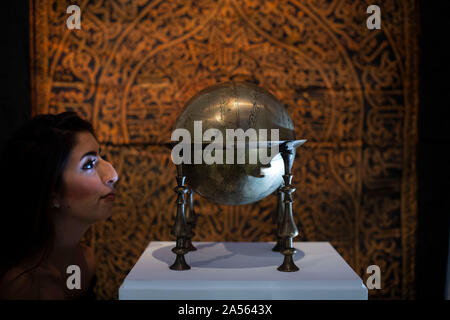 A staff member looks at a Safavid brass celestrial globe, made in Persia in 17th century (est. £80,000-120,000) in front of a rare Maghribi gilt-metal thread embroidered curtain, made in North Africa in 17th century (est. £50,000-70,000) during the preview of Middle Eastern Art Week at Sotheby's in London, ahead of their sale by auction on 22 and 23 October. PA Photo. Picture date: Friday October 18, 2019. Highlights of the sale will include a sumptuous leaf from the mythic 9th/10th century blue & gold Qu'ran, a dynamic painting of a great Indian epic wedding procession, jewellery including a
