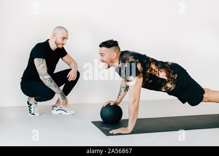 Fitness instructor observing man doing plank with medicine ball in studio Stock Photo