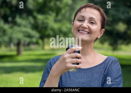 Portrait of middle aged woman holding a soft drink can and drinking soda, coke or beer outdoor in the park in a sunny summer day. Stock Photo