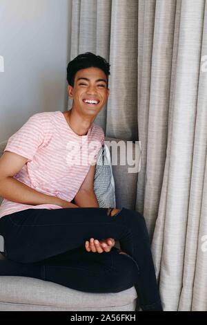 Young man sitting on bedroom chair laughing, portrait Stock Photo