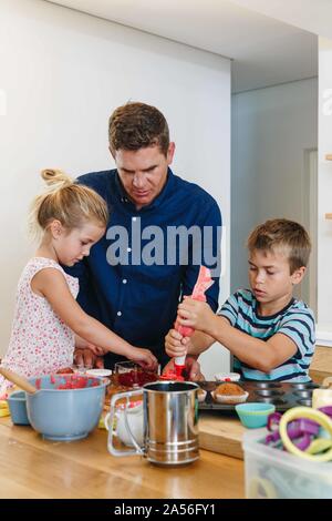 Father and children decorating cupcakes with frosting in kitchen Stock Photo