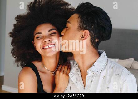 Happy young woman being kissed on cheek by boyfriend in bedroom, head and shoulders Stock Photo