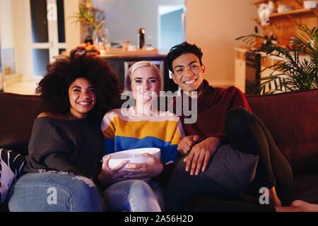 Young man and young female friends watching tv sitting on living room sofa with bowl of popcorn Stock Photo