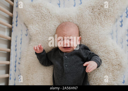 Baby boy crying while lying on sheepskin rug in crib, overhead view Stock Photo