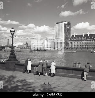 1960s, historical, people standing on the Victoria Embankment leaning on the pavement barrier looking over at the river Thames, towards the Southbank, London, England, UK. On the south side of the river, County Hall, the home of the GLC from 1965 to 1986, a new modern tower block under construction and on the far left, the Shot Tower beside the Royal Festival Hall. Stock Photo