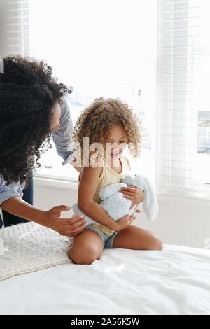 Woman talking to little girl cradling soft toy on bed Stock Photo