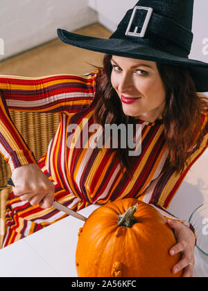 Beautiful young woman wearing witch's black hat  in her kitchen cutting a pumpkin for Halloween. Stock Photo