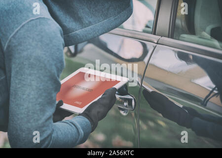 Hooded thief tries to break the car's security systems with tablet. Hacking modern car concept Stock Photo