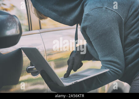 Hooded thief tries to break the car's security systems with laptop. Hacking modern car concept Stock Photo