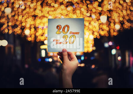 Hand holds a paper sheet with 2020 happy new year text message in the middle of a crowded night street with different Christmas lights and decorations Stock Photo