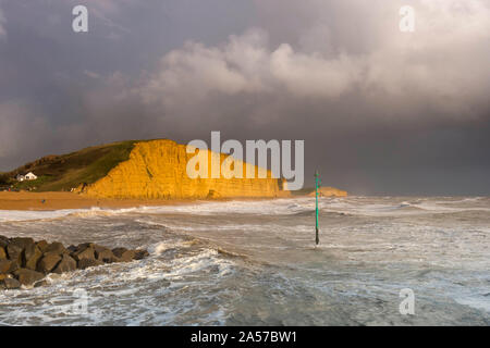 West Bay, Dorset, UK.  18th October 2019. UK Weather.  View of the East Cliff glowing a golden orange as dark stormy shower clouds clear from West Bay in Dorset after a day of sunshine and heavy showers. Picture Credit: Graham Hunt/Alamy Live News Stock Photo