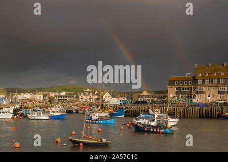 West Bay, Dorset, UK.  18th October 2019. UK Weather.  A rainbow rises above the harbour as dark stormy shower clouds clear from West Bay in Dorset after a day of sunshine and heavy showers. Picture Credit: Graham Hunt/Alamy Live News Stock Photo