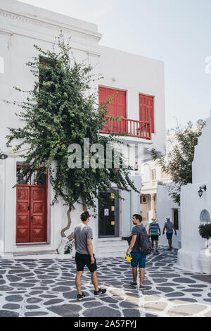 Mykonos Town, Greece - September 20, 2019: People walking past red door and shutters building on a street in Hora (also known as Mykonos Town), the is Stock Photo